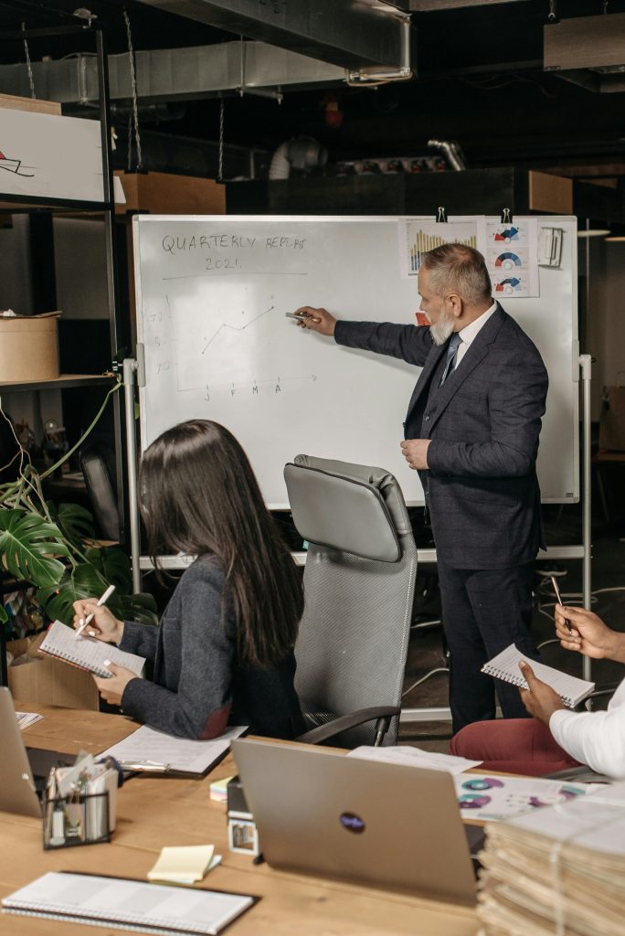 Corporate meeting scene with a presenter analyzing quarterly returns on a whiteboard in an office setting.